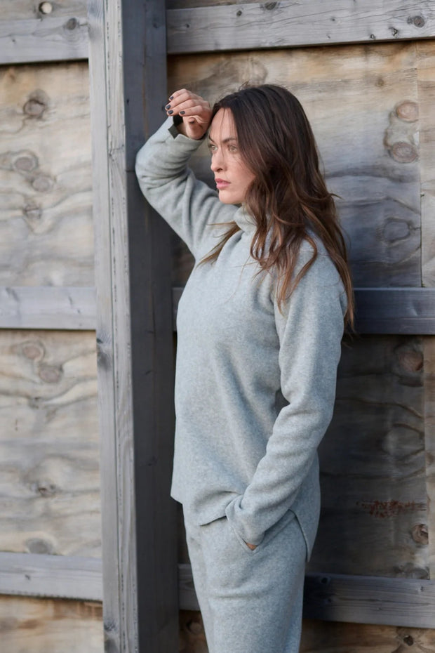 Woman in a light grey outfit leaning against a wooden wall.