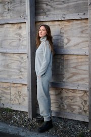 Woman in a light grey outfit standing against a wooden wall.