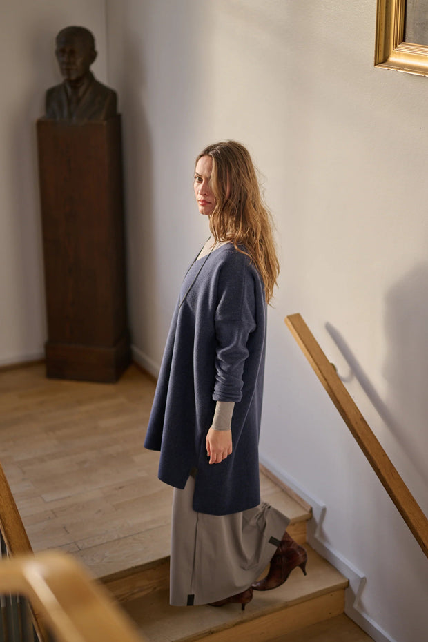 Woman in a blue tunic and gray dress standing on a staircase in an indoor setting.