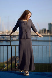 Woman in a long dark navy dress standing by a waterfront with boats in the background