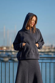Woman wearing a dark hoodie and pants standing by a waterfront with boats in the background.