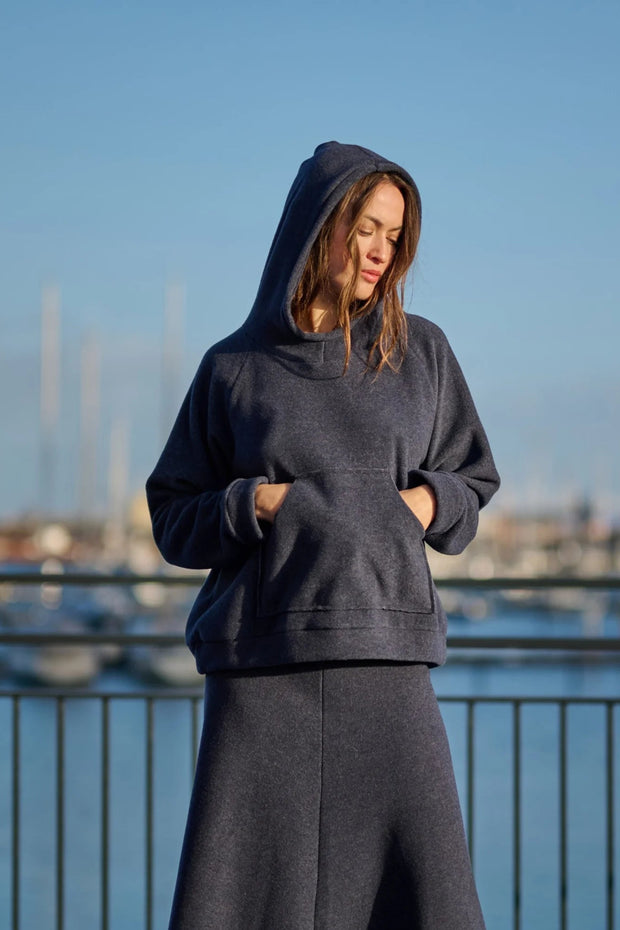 Woman wearing a dark hoodie and pants standing by a waterfront with boats in the background.