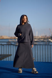 Person wearing a dark hoodie and skirt standing on a blue surface with a marina and skyline in the background.