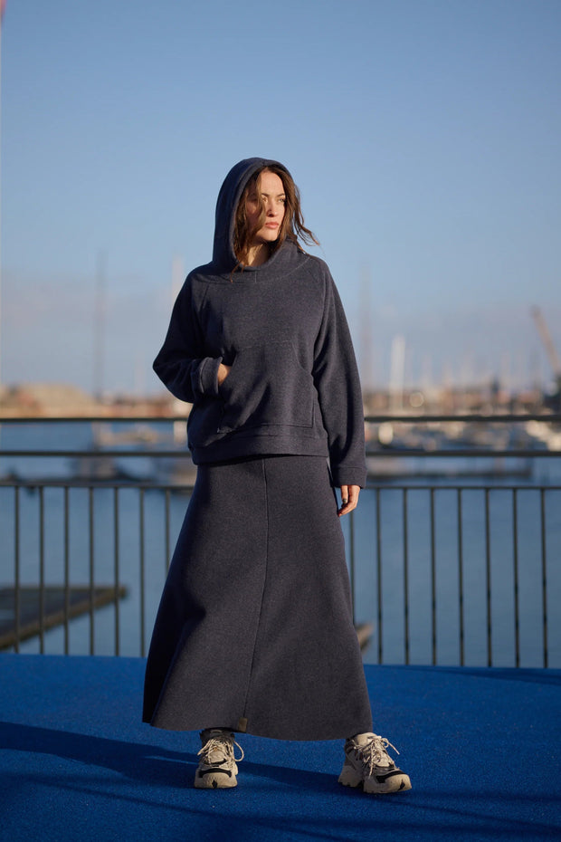 Person wearing a dark hoodie and skirt standing on a blue surface with a marina and skyline in the background.