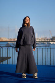 Person wearing a dark hoodie and skirt standing by a waterfront with a clear blue sky.
