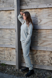 Woman in a light gray outfit leaning against a wooden wall.