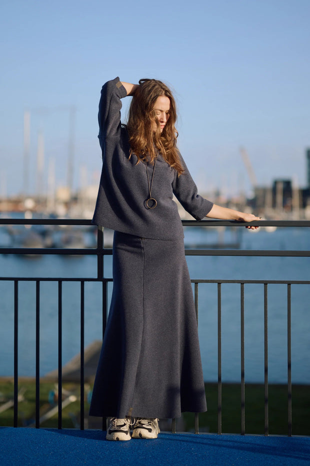 Woman in a long navy top and skirt standing by a waterfront with boats and buildings in the background.