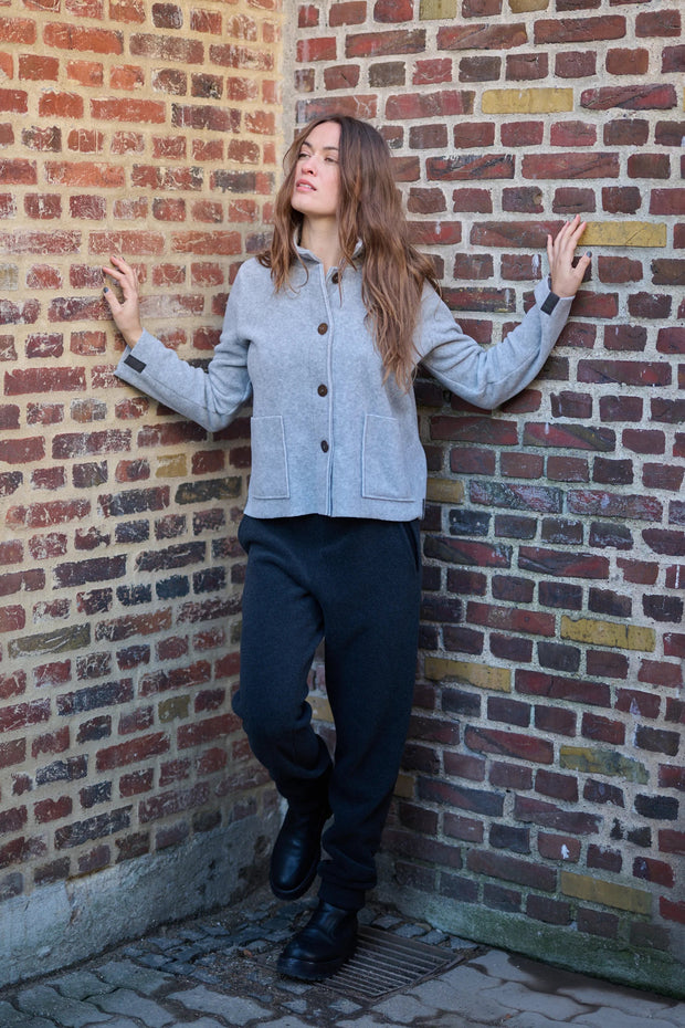 Woman leaning against a brick wall wearing a gray jacket and dark pants.