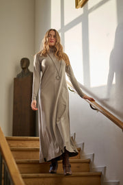 Woman in a long beige dress standing on a staircase with sunlight streaming in.